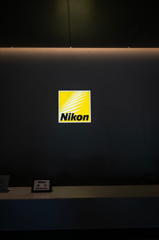 A Nikon logo illuminated on a dark wall above a reception desk. The logo is bright yellow with the word "Nikon" in black text, featuring a design of diagonal lines. The reception area is dimly lit.