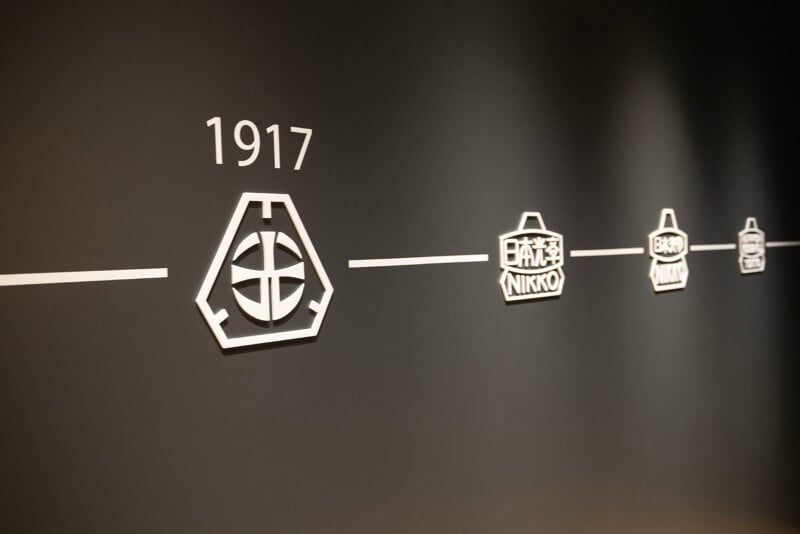 A timeline on a dark wall showing the evolution of a logo design. It starts with a 1917 emblem and progresses with different versions along the line, including text in Japanese characters and the word "NIKKO.