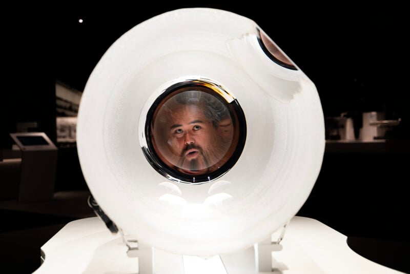 A man looks through a large, circular, transparent tube. His face is centered within the tube, creating a distorted view. The background is dark with blurred objects and screens.