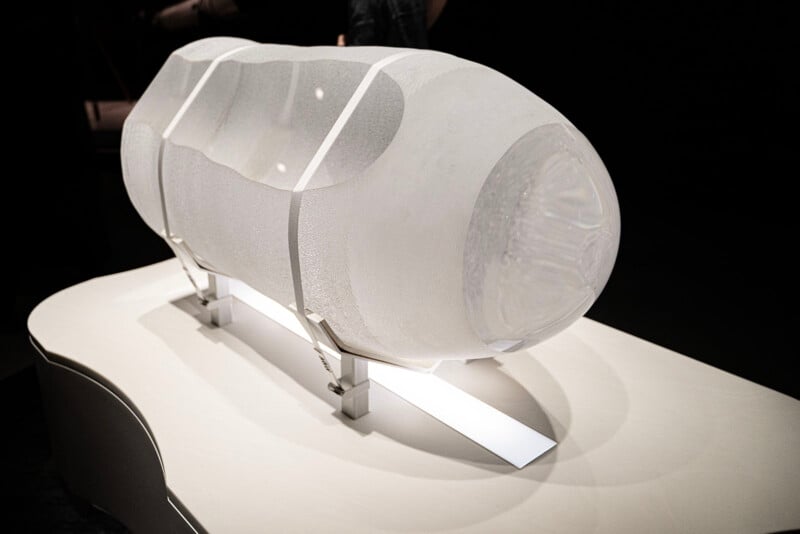 An abstract, translucent sculpture with a cylindrical shape is displayed on a white platform in a dimly lit environment. The sculpture has a textured surface and is supported by metal brackets.