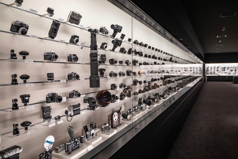 A vast display of cameras and lenses is showcased in a well-lit museum exhibit. The items are arranged on multiple glass shelves along a long corridor, highlighting the evolution and variety of photographic equipment.