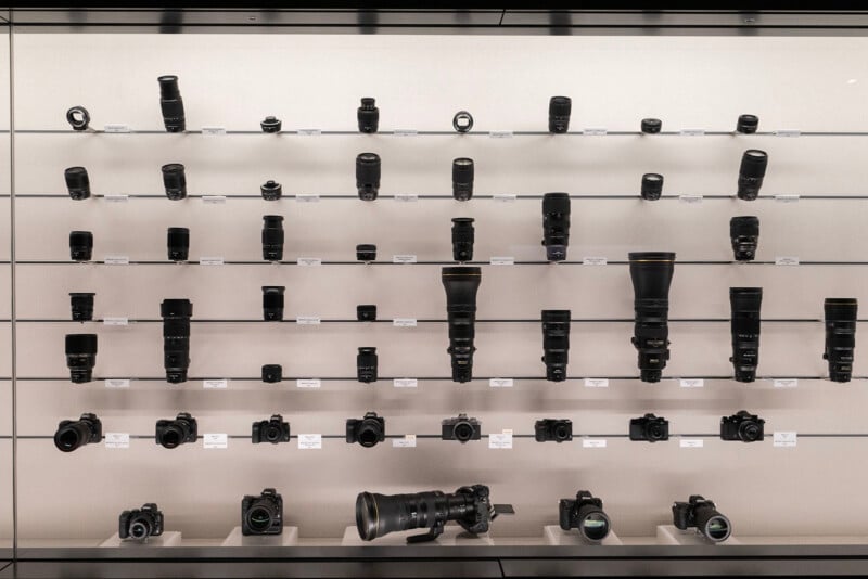 A display of various camera lenses and bodies arranged on shelves. The lenses vary in size and type, showing different focal lengths and designs. The shelves are well-lit, highlighting the equipment against a neutral background.