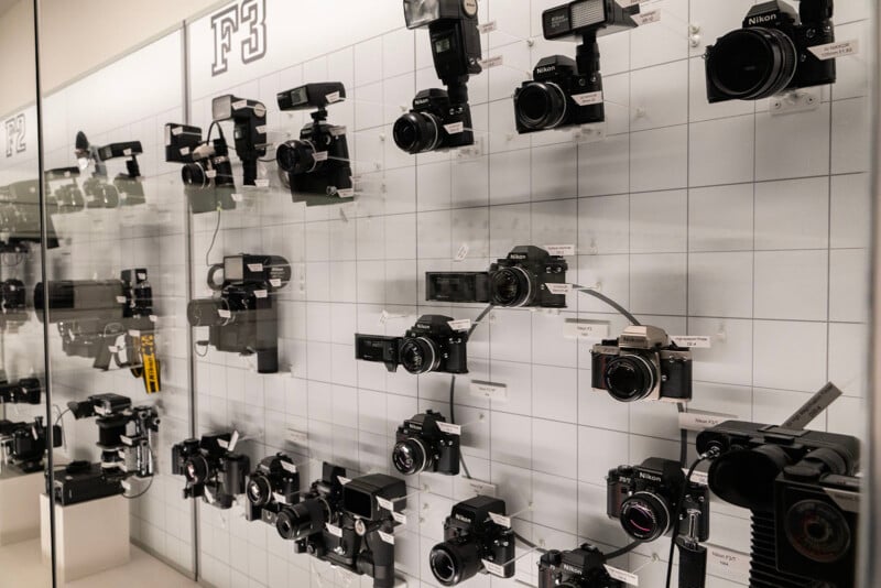 A display wall featuring a collection of vintage cameras, including various models with lenses and attachments, showcased against a grid-patterned background. Some cameras have labels underneath.