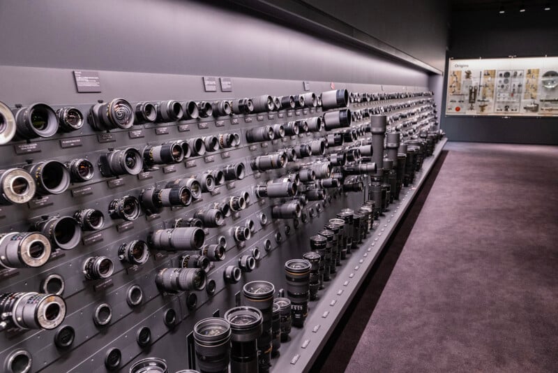 A long wall display showcases a wide array of camera lenses in various sizes and types. The lenses are arranged neatly on shelves, each section labeled. The room has a dark interior, and a glass display is visible at the far end.