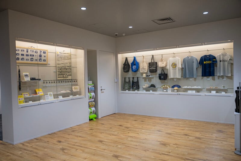 A store display showcasing various merchandise. On the left, shelves with books, postcards, and small items. On the right, t-shirts and bags hang above glass cases displaying more products. The floor is wooden, and the walls are light-colored.