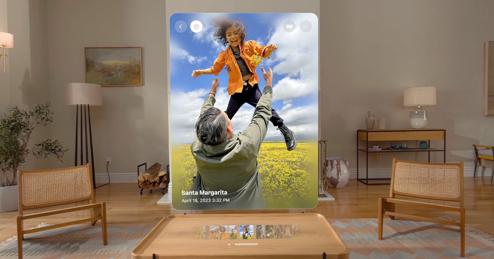 A person is sitting in a living room, viewing a large digital photo of an adult joyfully lifting a child in a field of yellow flowers. Text on the photo reads "Santa Margarita, April 18, 2023, 3:42 PM." The room has modern furniture and decor.