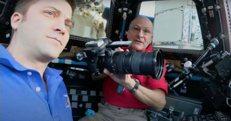  one holds a large camera, wearing a red shirt, while the other, in a blue shirt, looks toward it. Background details include control panels and spacecraft interior.