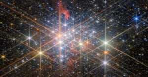 A vibrant star cluster set against a dark space background. Numerous stars shine brightly, with diffraction spikes creating starburst effects. Wisps of red nebula gas are interspersed throughout, adding texture and depth to the celestial scene.