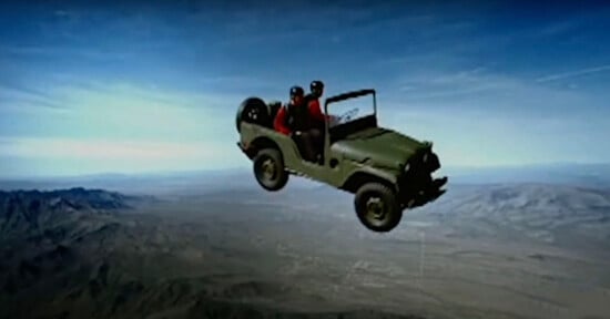 An off-road vehicle with two passengers appears to be airborne against a wide desert landscape and a blue sky.