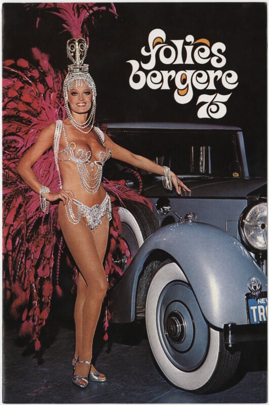 A performer in an elaborate costume adorned with feathers and sparkling jewels poses next to a vintage car. The text "Folies Bergere" appears at the top of the image.