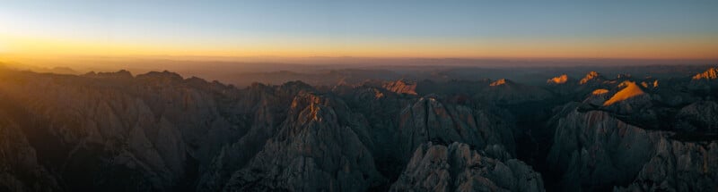 A panoramic view of rugged mountains at sunset. The warm glow of the sun casts golden highlights over the peaks, creating a dramatic contrast with the deep shadows in the valleys. The horizon stretches under a clear sky, fading into the distance.