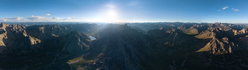 A panoramic view of a mountainous landscape at sunset, with rugged peaks casting long shadows over a valley that includes a small lake. The sky is clear, with the sun positioned low on the horizon, creating a serene and majestic atmosphere.