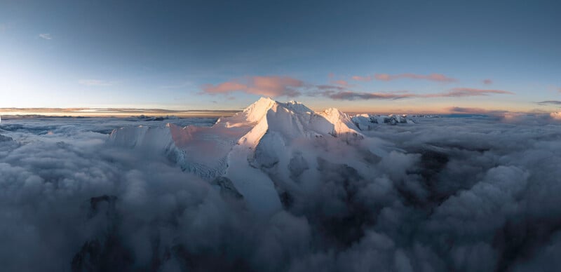 A majestic snow-capped mountain peak bathed in golden light rises above a sea of fluffy clouds. The expansive sky is tinged with hues of pink and blue as the sun sets, creating a serene and breathtaking scene.