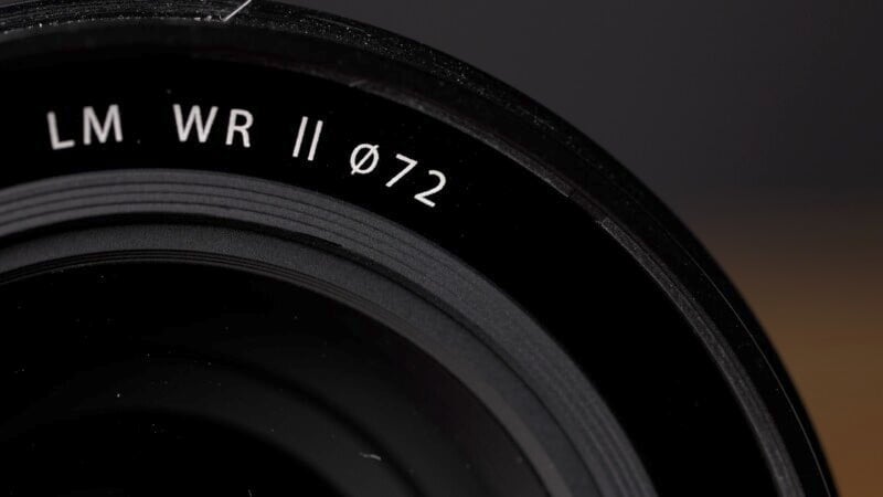 Close-up of a camera lens with inscriptions "LM WR II Ø72" visible on the outer rim. The image focuses on the lens's textured surface, against a dark background.