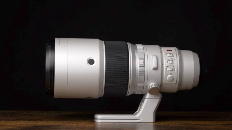 A large, white camera telephoto lens with various controls and a tripod mount sits on a wooden surface against a black background. The lens is positioned horizontally, showcasing its buttons and switches.