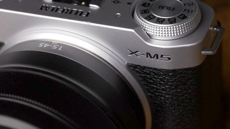 Close-up of a Fujifilm X-M5 camera showing the textured grip, lens, and part of the silver top plate with a control dial.