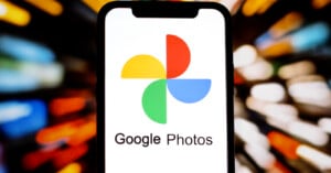 A smartphone displays the Google Photos logo on its screen, featuring a multicolored pinwheel icon. The background is blurred with streaks of orange, yellow, and white lights, creating a dynamic effect.