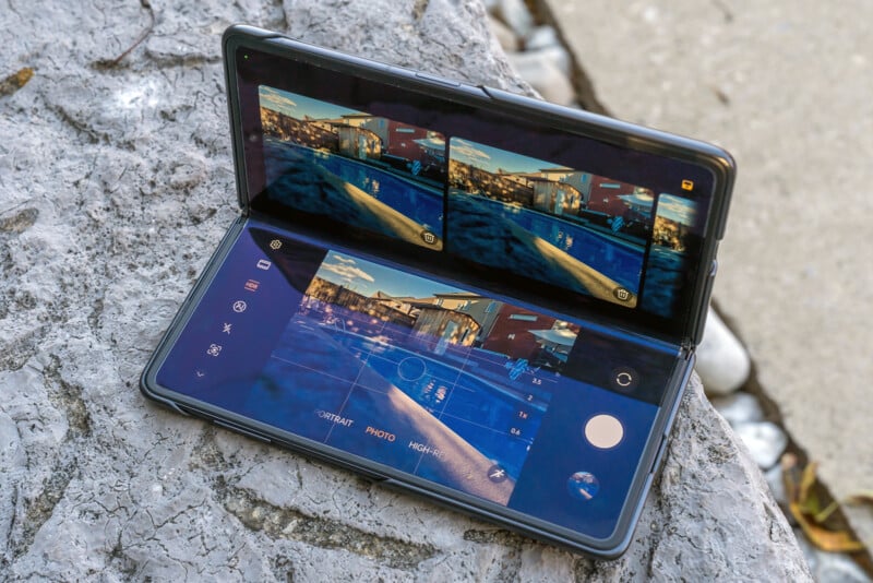 A foldable smartphone is placed on a stone surface, displaying a dual-screen camera app. The screens show a cityscape with buildings and a waterfront, with camera controls visible below the images.