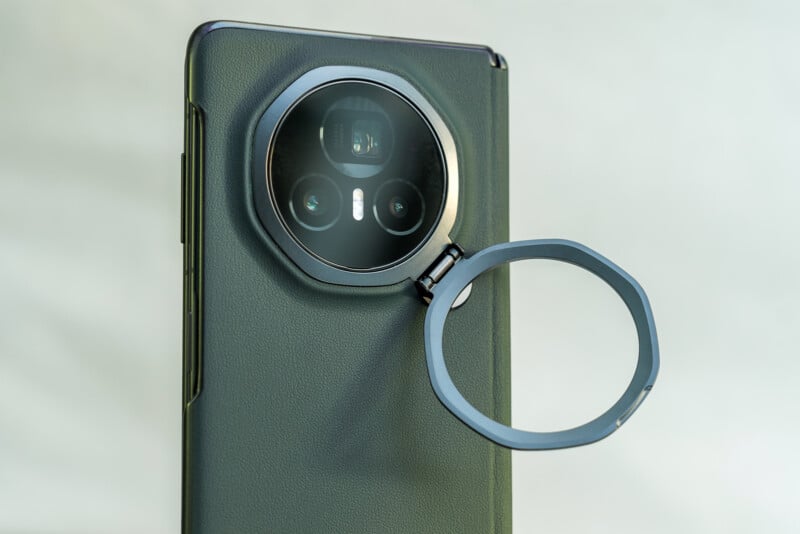 A smartphone with a circular camera module featuring three lenses. A removable ring attachment is partially detached, showing its functionality. The phone has a textured dark-colored back and a sleek design against a light background.