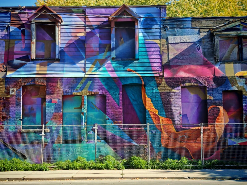 A colorful mural on a building wall features abstract geometric shapes in vibrant shades of purple, blue, orange, and teal. The mural covers windows and doors, with a fence and greenery in front of the building.