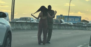 Two people are walking on a highway between lanes of slow-moving traffic. The person on the left is flipping their long hair, and the person on the right is wearing a hoodie. A billboard in the background says "SUN'S UP.