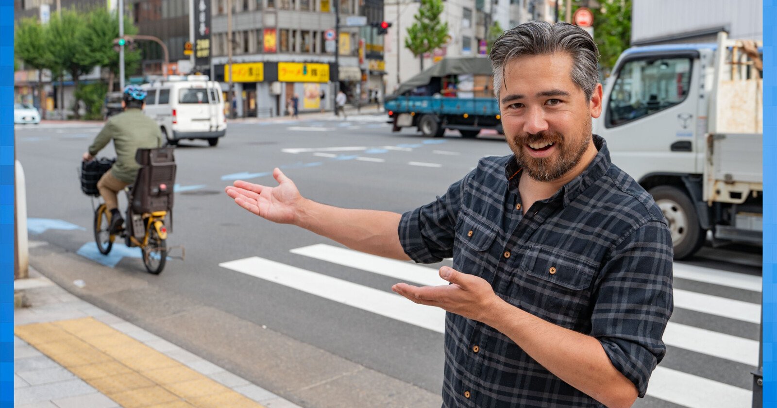 A smiling man in a plaid shirt gestures toward a bustling city street with vehicles and a cyclist. The scene features a crosswalk, buildings, and a bright, sunny day.