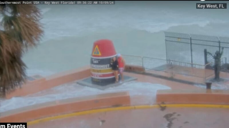 A couple stands by the Southernmost Point buoy in Key West, Florida, as waves crash over the seawall. The weather appears stormy with whitecaps visible in the ocean. Palm tree leaves partially frame the left side of the image.