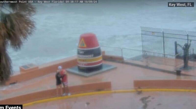 A blurry photo of the Southernmost Point buoy in Key West, Florida, during heavy rain. Two people stand near the buoy, with strong waves crashing in the background. A palm tree is visible on the left side.