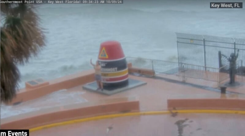 A person stands near the Southernmost Point marker in Key West, Florida, during a storm. The ocean is rough, and waves are crashing over the barrier onto the wet pavement. Overcast skies dominate the background as palm trees sway in the wind.