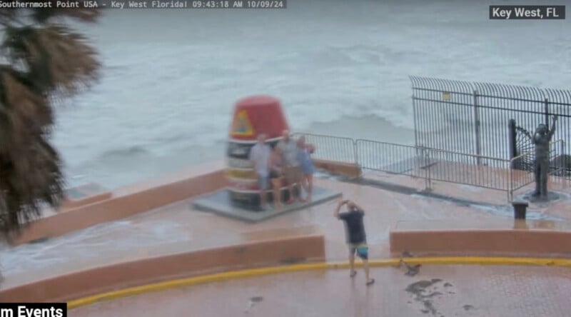 A group of people stands near the Southernmost Point buoy in Key West, Florida, as waves crash over the seawall. The ocean is rough and the sky is overcast, indicating stormy weather.