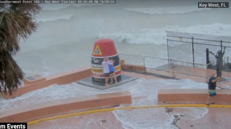 People stand at the Southernmost Point buoy in Key West, Florida, surrounded by rising ocean waves. The water covers parts of the walkway, and the sky appears overcast.