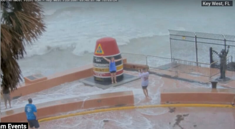 A stormy ocean scene in Key West, FL, with people standing near the Southernmost Point buoy. Water waves splash over the barrier onto the viewing area, and one person is posing with arms raised next to the buoy. Cloudy sky and rough seas in the background.