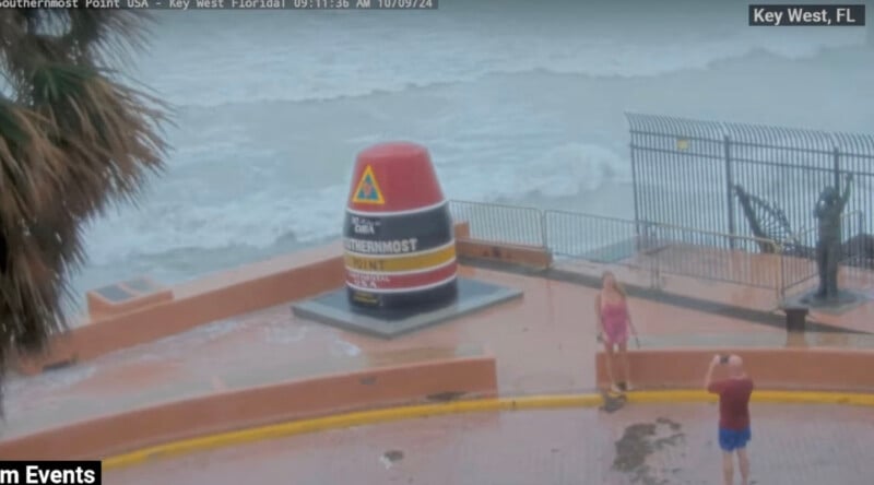 A seaside view in Key West, FL, showing the Southernmost Point buoy, surrounded by waves. Two people stand nearby, one taking a photo of the other amidst misty weather. A palm tree is partially visible on the left.