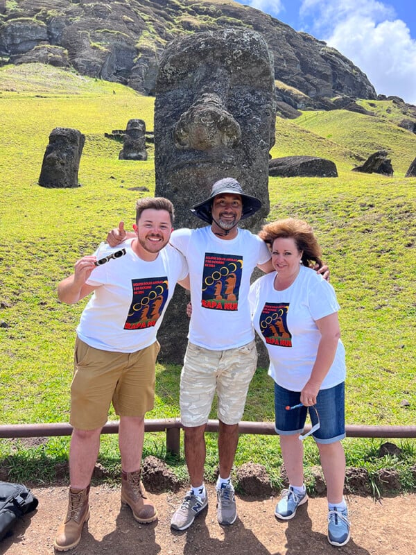 Three people wearing matching white T-shirts stand together, smiling in front of a large Moai statue on a grassy hill. The sky is blue with scattered clouds.