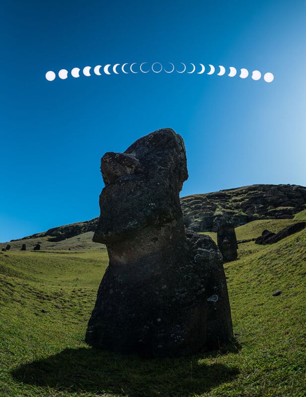 A monolithic stone statue stands on a grassy hillside under a clear blue sky. Above the statue, there is a sequence of moon phases depicted in a curved line.