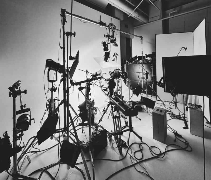 A black and white photo of a cluttered photography studio filled with various lighting equipment and stands. Circular reflectors, cables, and tripods are scattered around, creating a busy and intricate setup, with a bright backdrop in the center.