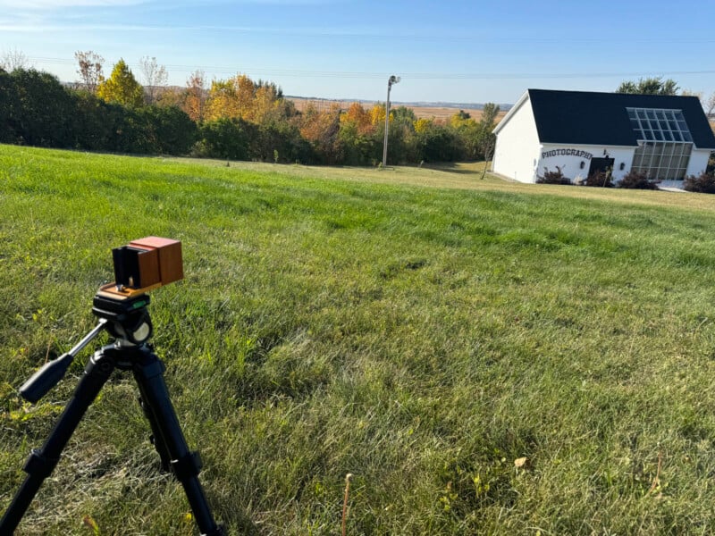 A grassy field with a vintage camera on a tripod in the foreground. In the background, there's a small white building with a solar panel roof and "PhotoPills" written on it, surrounded by trees under a clear blue sky.