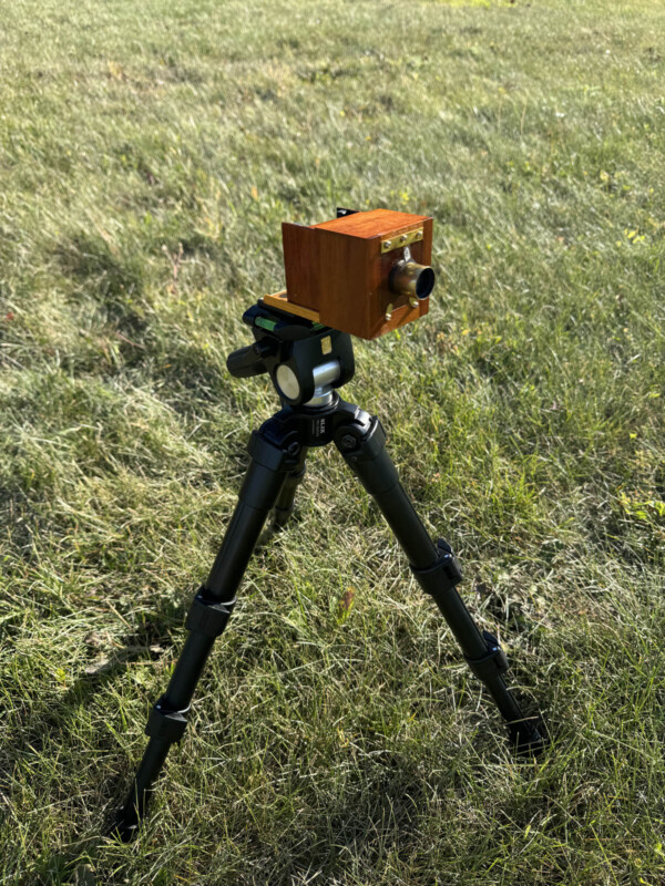 A wooden pinhole camera mounted on a black tripod stands on green grass in a field. The sunlight casts soft shadows on the ground, highlighting the camera's rustic design against the natural background.