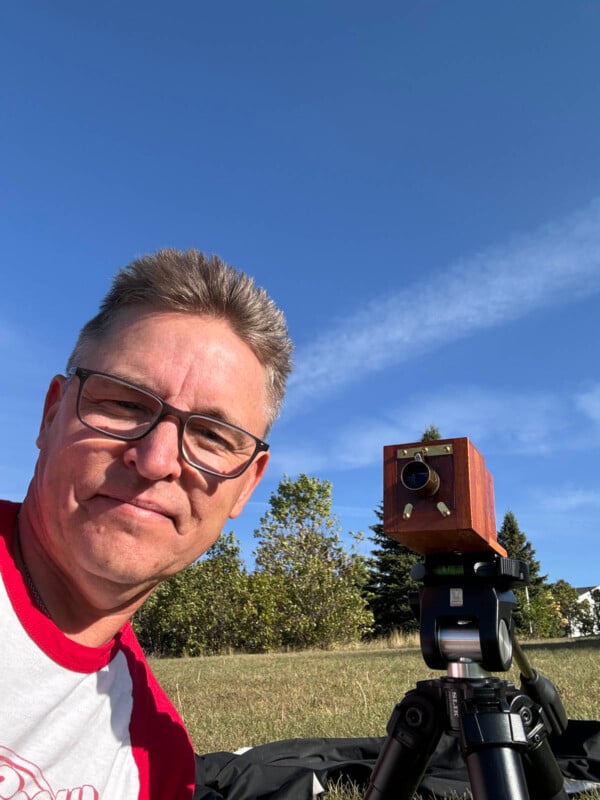 A person wearing glasses and a red and white shirt smiles at the camera. They are outdoors, with trees and a clear blue sky in the background. A wooden camera sits on a tripod beside them.