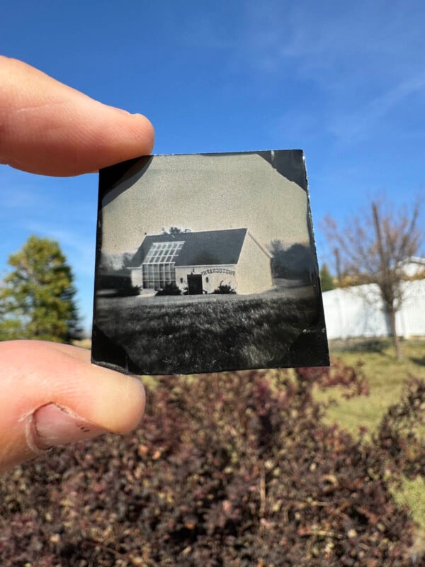 A hand holds a small vintage photograph negative against a clear blue sky. The image within shows a building with a gabled roof, surrounded by grass, trees, and a white fence in the background.