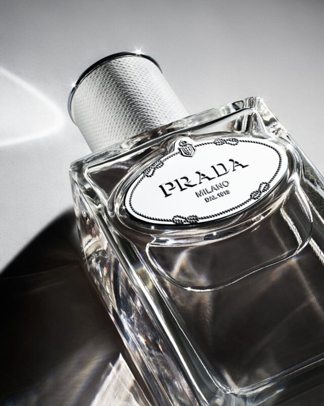 A close-up of a clear, elegant Prada perfume bottle with a textured silver cap. The label reads "Prada Milano" and reflects light, casting a subtle shadow on a light surface.