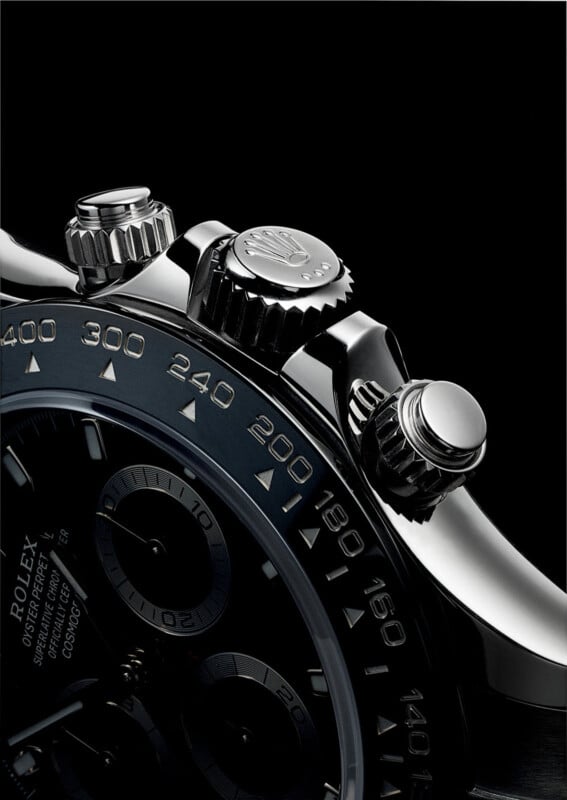 Close-up of a luxury wristwatch with a dark face and shiny metal details. The focus is on the side controls and bezel featuring engraved numbers. The sleek design and polished surface reflect light, emphasizing the watch's craftsmanship.