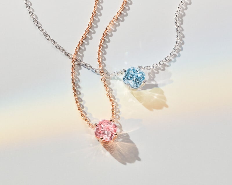 Two delicate necklaces are displayed on a light surface. Each features a round gemstone—one pink on a rose gold chain, the other blue on a silver chain. A soft rainbow reflection adds a subtle, colorful touch to the composition.