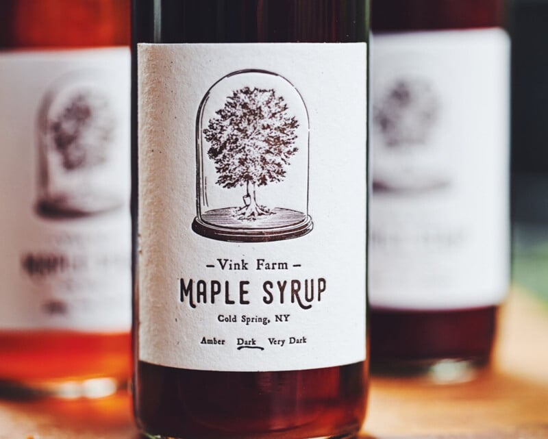 Close-up of a bottle of maple syrup from Vink Farm, labeled "Cold Spring, NY." The label features an illustration of a tree under a glass dome and indicates the syrup is available in Amber, Dark, and Very Dark varieties.