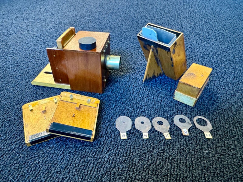 A vintage wooden camera set with several components on a blue carpet. The setup includes a camera box with a lens, film holders, wooden plates, and numbered metal keys.