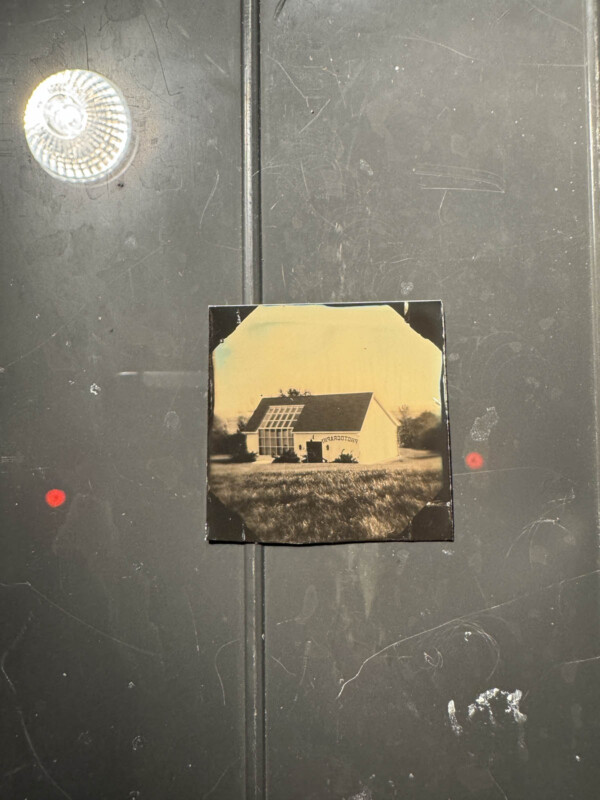 A black and white photograph of a small house with a sign that reads "Laboratory" on it. The photo is mounted on a dark surface with two red dots and a round reflective light visible.