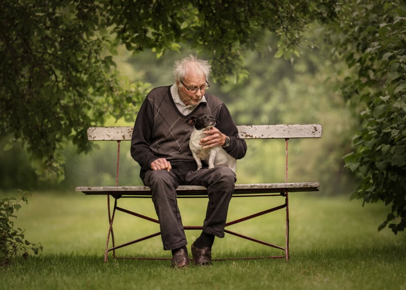 An elderly man sitting on a wooden bench outdoors, holding a small dog. He is surrounded by lush green trees and grass, creating a peaceful and serene setting.