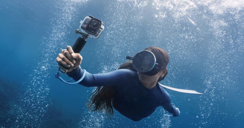 A person wearing a wetsuit and snorkel mask is underwater, holding a camera on a stick. Bubbles surround them in the clear blue water, suggesting movement.