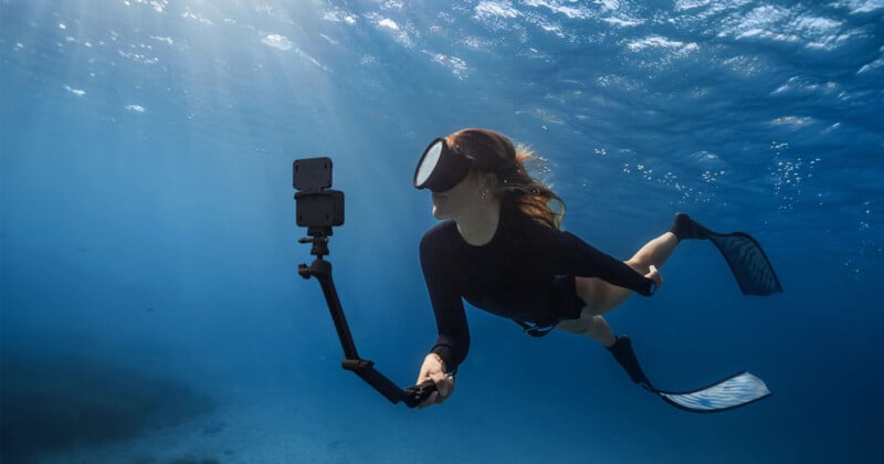 A person wearing a snorkel mask and fins swims underwater while holding an extended camera. Sunlight filters through the water, casting rays above. The scene captures a serene aquatic environment with a clear blue backdrop.