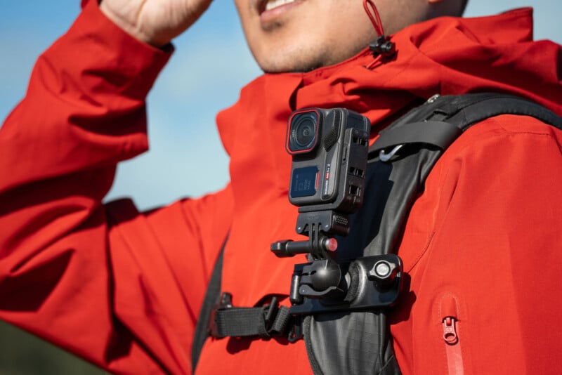 A person wearing a red jacket and black backpack with a mounted action camera on the shoulder strap. The individual is adjusting the backpack or saluting, with a blurred outdoor background.